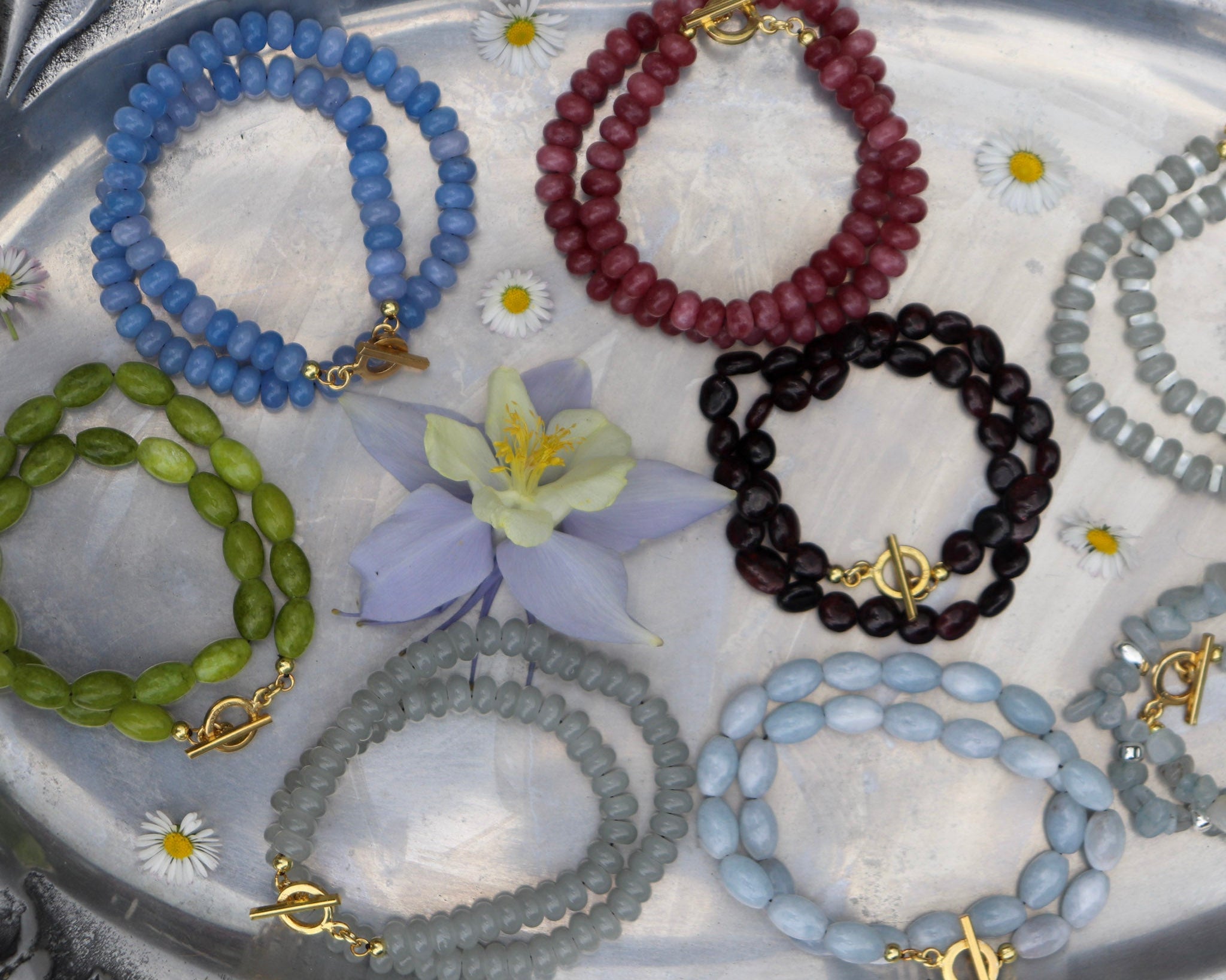 Collection of beaded necklaces in various colors on a textured surface with flowers.